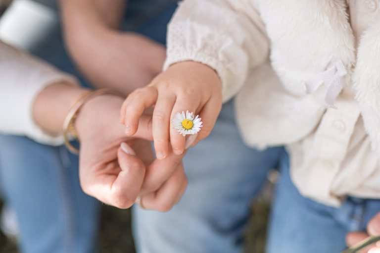 Photographie de détail des mains d'une maman et de sa fille de 2 ans. Elle tiennent une pâquerette.